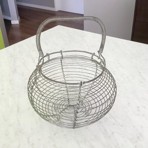 May include: A round wire basket constructed from thin silver-toned metal strands. The basket features a wide, open top and a singular, arched handle made of coiled wire that is attached to the rim with metal loops for a secure, flexible grip.