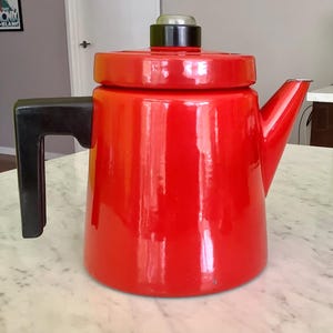 May include: A vibrant red enamel coffee pot with a black handle and lid. The pot has a classic design with a spout for pouring. The lid features a silver-colored top. The coffee pot is a kitchenware item.