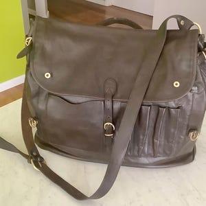 May include: A dark brown leather messenger bag with a flap closure, shoulder strap, and multiple exterior pockets. The bag features brass-colored hardware and a top handle. The bag is sitting on a white surface.