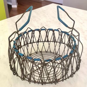 May include: An antique wire basket with a collapsible design. The basket has a circular shape with a dark metal frame and blue accents on the rim and handles. The handles are also blue.