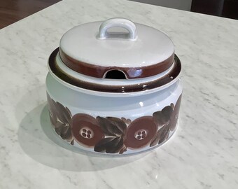 May include: A white ceramic serving dish with a lid. The dish has a brown floral pattern around the base and a brown band around the rim. The lid has a white handle and a brown band.