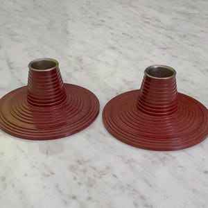 May include: Two red candle holders with a layered design. Each holder has a circular base and a tall, conical centre piece.