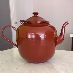 May include: A glossy, deep rust-colored teapot with a rounded body, a curved handle, and a matching lid featuring a small knob. The teapot has a short, tapered spout and a smooth, reflective finish, showcasing a simple and functional design.