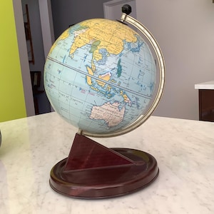 May include: A vintage-style world globe featuring a pale blue ocean and yellow landmasses with printed geographical labels. The globe is mounted on a metallic meridian ring and supported by a dark brown, triangular-shaped wooden base stand.