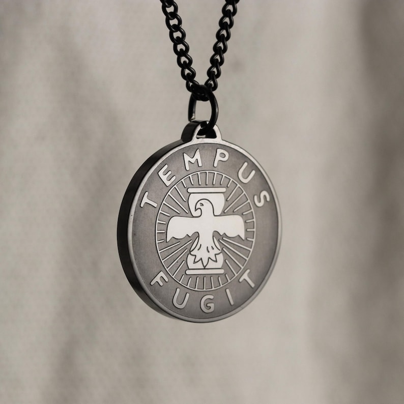 May include: A black chain necklace with a round silver pendant. The pendant features an eagle with outstretched wings, a sundial, and the words "TEMPUS FUGIT" around the edge.
