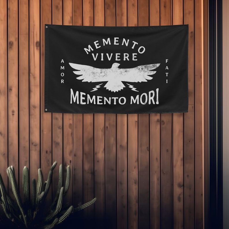 May include: Black flag with white text and a distressed eagle graphic. The flag reads "MEMENTO VIVERE" above the eagle, and "MEMENTO MORI" below. The words "AMOR" and "FATI" are on the left and right sides of the eagle.