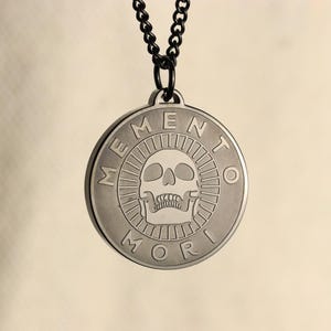 May include: A silver pendant necklace with a black chain. The pendant is round and features a skull with the words "Memento Mori" around the edge.
