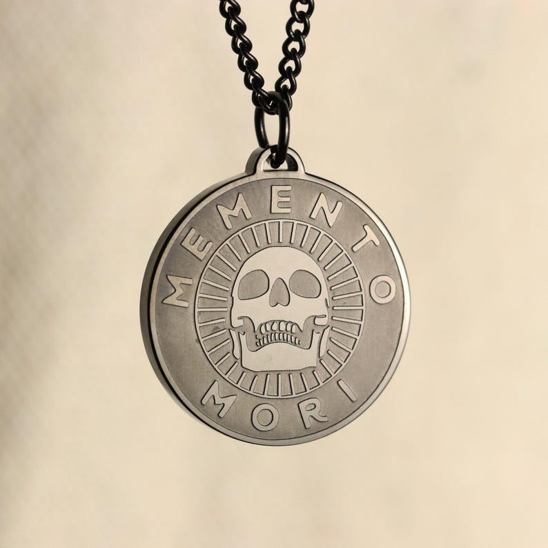 May include: A silver pendant necklace with a black chain. The pendant is round and features a skull with the words "Memento Mori" around the edge.