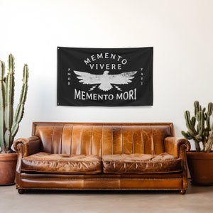 May include: Black wall tapestry with white text and a bird graphic. The text reads "Memento Vivere, Memento Mori, Amor Fati." The tapestry is hanging above a brown leather couch. Two potted cacti are on either side of the couch.