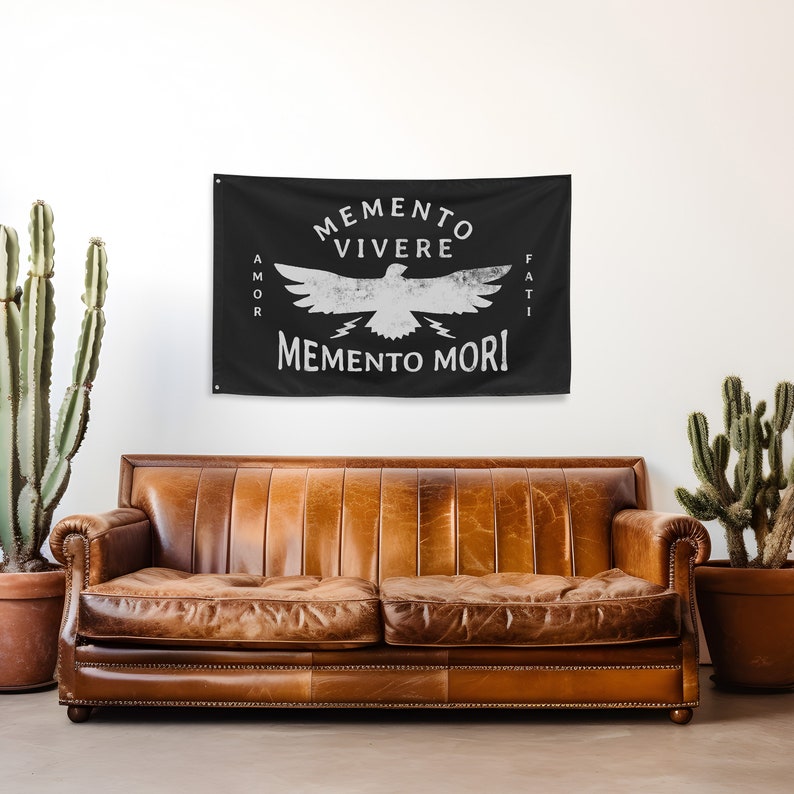 May include: Black wall tapestry with white text and a bird graphic. The text reads "Memento Vivere, Memento Mori, Amor Fati." The tapestry is hanging above a brown leather couch. Two potted cacti are on either side of the couch.