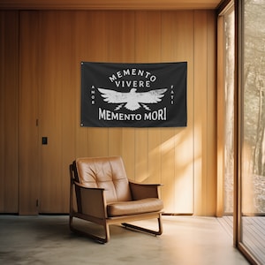 May include: A black flag with white text and a distressed eagle design. The flag reads "Memento Vivere" above the eagle, and "Memento Mori" below. The words "Amor" and "Fati" are on the left and right sides. The flag is hanging on a wooden wall.