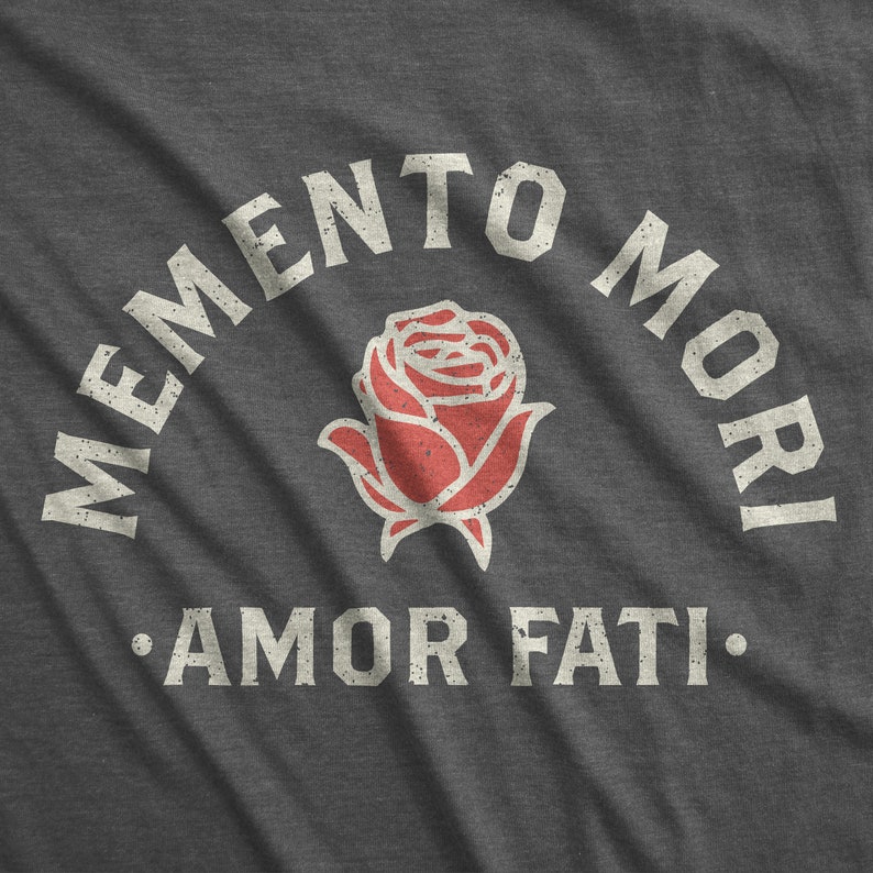 Op de afbeelding: Donkergrijze t-shirt met een distressed design. Het ontwerp toont de woorden "MEMENTO MORI" boven een rode rozenillustratie, met "AMOR FATI" eronder. De tekst en de roos hebben een vintage, versleten uitstraling.