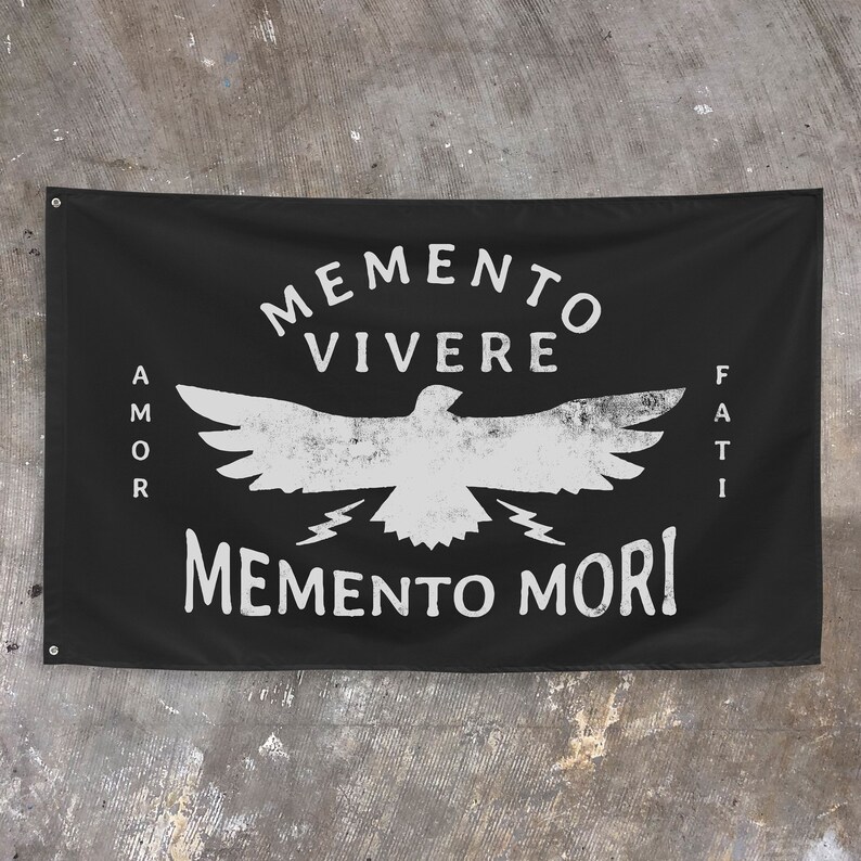 May include: Black banner with white text and a distressed bird graphic. The banner reads "MEMENTO VIVERE" above the bird, "MEMENTO MORI" below, and "AMOR FATI" on the sides. The banner is hanging on a concrete wall.