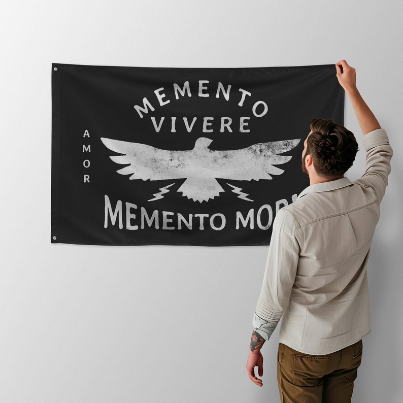 May include: Black rectangular flag with white distressed text and a bird graphic. The text reads "MEMENTO VIVERE" above the bird, "MEMENTO MORI" below, and "AMOR" on the left side. The flag is held up by a person.