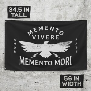 May include: Black flag with white distressed text and graphic. The flag reads "Memento Vivere" above a bird graphic, with "Amor" on the left and "Fati" on the right. Below the bird is "Memento Mori". The flag is 34.5 inches tall and 56 inches wide.