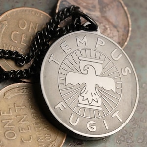 May include: A silver-toned pendant necklace with a circular design featuring a stylized eagle and the Latin phrase "Tempus Fugit" (Time Flies). The necklace is on a black chain and is resting on top of several US pennies.