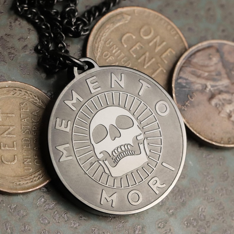 May include: A silver-toned pendant necklace with a black chain. The pendant features a skull with a white face and the words "Memento Mori" around the edge.