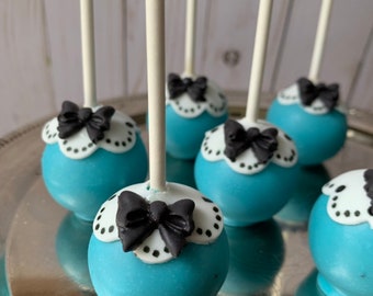 May include: A close-up of six blue and white cake pops with black bows. The cake pops are on a silver tray and have white sticks.