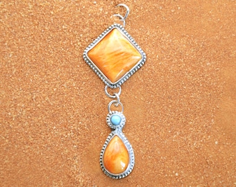 May include: A silver pendant with a teardrop-shaped orange stone and a square orange stone. The pendant is on a brown background.