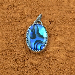 Peut inclure: Un pendentif en argent avec une incrustation de nacre bleue et noire. Le pendentif est de forme ovale et a un bord festonné.