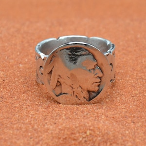 May include: A silver-toned metal ring featuring a circular centerpiece embossed with the profile of a person wearing a feathered headdress. The band of the ring displays a repeating decorative pattern along its outer surface with a metallic finish.