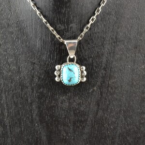 Puede incluir: Un collar de cadena de plata con un colgante turquesa. El colgante es de forma cuadrada con un borde de plata y una piedra turquesa en el centro.