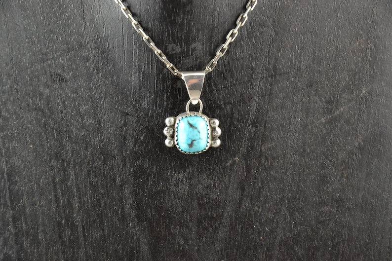 Puede incluir: Un collar de cadena de plata con un colgante turquesa. El colgante es de forma cuadrada con un borde de plata y una piedra turquesa en el centro.
