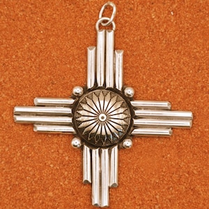 Peut inclure: Un pendentif en argent en forme de symbole traditionnel amérindien, le symbole du soleil Zia. Le symbole est composé de plusieurs barres d'argent qui se croisent au centre, formant une croix. Le centre de la croix présente un motif rond de couleur or avec un motif de soleil.