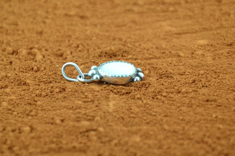 Puede incluir: Un colgante de plata con una piedra ovalada blanca y un borde decorativo. El colgante est&aacute; sobre una superficie marr&oacute;n.