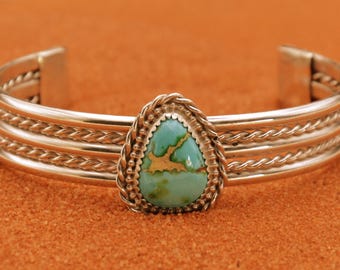 May include: Silver cuff bracelet featuring a turquoise stone. The bracelet has a braided design and a teardrop-shaped turquoise stone in the center. The turquoise stone has a mix of green and brown colors.