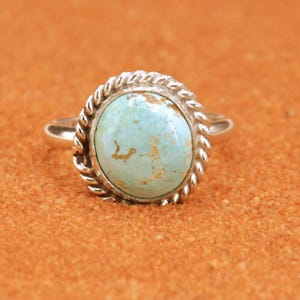 Puede incluir: Anillo de plata con una gran piedra turquesa ovalada. La piedra es de color azul verdoso pálido con inclusiones marrones. El anillo tiene un diseño de cuerda trenzada alrededor de la piedra y una banda sencilla.