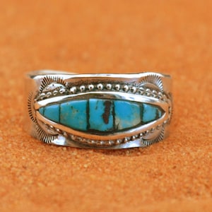 May include: A silver ring with a rectangular turquoise stone inlay. The ring has a detailed design with beaded and stamped patterns. The turquoise is a vibrant blue-green color, set in a silver band. The ring is displayed on an orange surface.