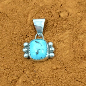 Puede incluir: Un colgante de turquesa con un engaste de plata y una anilla de plata. El colgante est&aacute; sobre un fondo marr&oacute;n.