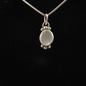 Puede incluir: Un collar de cadena de plata con un colgante que presenta una piedra ovalada blanca rodeada por un borde de cuentas de plata.