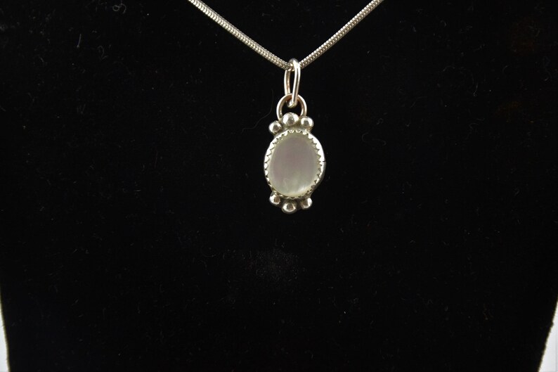 Puede incluir: Un collar de cadena de plata con un colgante que presenta una piedra ovalada blanca rodeada por un borde de cuentas de plata.