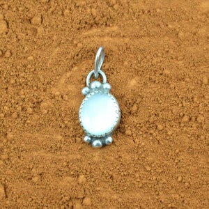 Puede incluir: Un colgante de plata con una piedra de &oacute;palo blanco y un borde de cuentas. El colgante est&aacute; sobre un fondo marr&oacute;n.