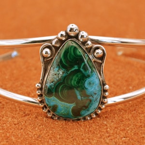 May include: A silver bracelet featuring a teardrop-shaped green and blue gemstone set in a silver bezel. The bracelet has a simple, elegant design.