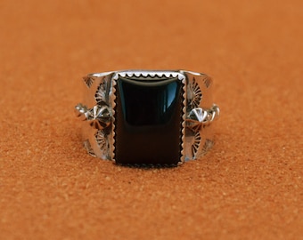 Könnte beinhalten: Ein silberner Ring mit einem großen, rechteckigen schwarzen Stein. Der Ring hat aufwendige geätzte Designs und dekorative Elemente am Band. Der Hintergrund ist eine strukturierte, orange-braune Oberfläche.