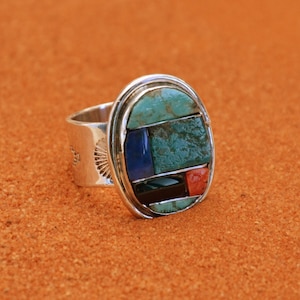 Può includere: Un anello d'argento con una pietra ovale incastonata al centro. La pietra è composta da più pezzi di turchese, lapislazzuli, corallo e onice nero.