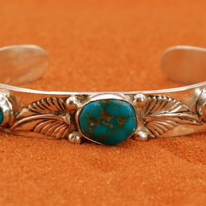 May include: A silver cuff bracelet featuring three oval turquoise stones with brown matrix patterns. The metal band is decorated with embossed leaf motifs and small silver spheres positioned between the stones and the leaf designs.