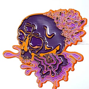 May include: A purple skull enamel pin with orange and purple accents. The skull has a crystal-like design on top and a dripping effect at the bottom, all set against a white background. The pin is a unique accessory.