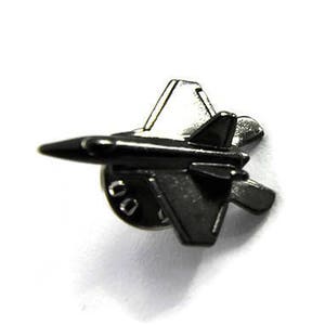 May include: A small, dark gray metal airplane pin. The pin features a detailed design of a jet fighter aircraft, with wings, tail, and a pointed nose. The pin is likely made of a metal alloy and has a shiny finish.