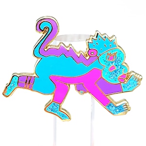 May include: A colorful enamel pin featuring a stylized monkey design. The pin has a gold outline and is filled with bright turquoise, purple, and pink enamel. Glitter accents the monkey's crown, hands, and feet. The monkey is in a playful pose.