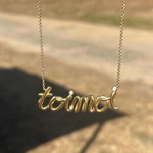 May include: A delicate gold-colored necklace with the name "toimoi" in cursive script. The necklace hangs against a blurred outdoor background of a dirt road, trees, and a clear blue sky. The chain is thin and the pendant is in a handwritten style.
