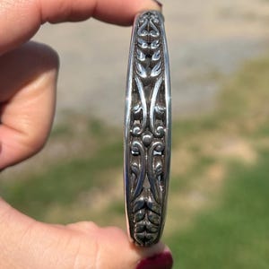 May include: A silver cuff bracelet featuring a continuous, raised, symmetrical relief pattern of stylized leaves and curved, vine-like elements. The metal has a polished finish with darkened, recessed areas that highlight the intricate, repeating design.