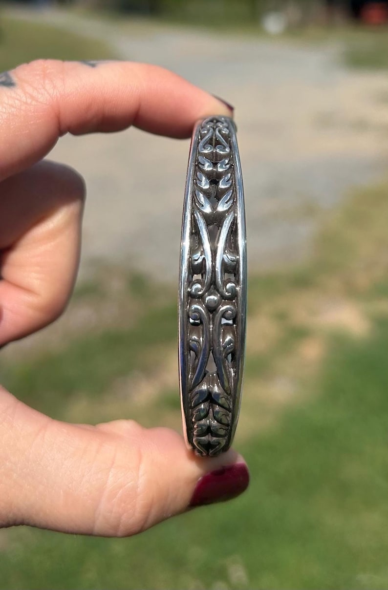 May include: A silver cuff bracelet featuring a continuous, raised, symmetrical relief pattern of stylized leaves and curved, vine-like elements. The metal has a polished finish with darkened, recessed areas that highlight the intricate, repeating design.