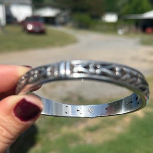 May include: A silver-toned metal bangle bracelet featuring an intricate, repeating openwork pattern along the outer surface. The interior of the band is smooth and polished, displaying a small, stamped heart-shaped hallmark logo centered inside.