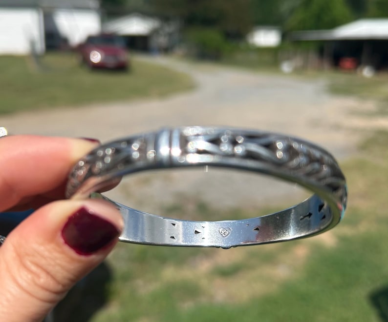 May include: A silver-toned metal bangle bracelet featuring an intricate, repeating openwork pattern along the outer surface. The interior of the band is smooth and polished, displaying a small, stamped heart-shaped hallmark logo centered inside.