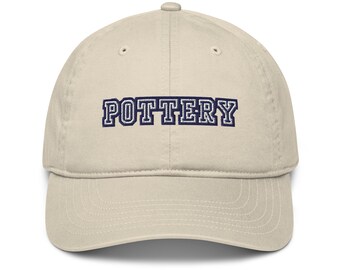Może przedstawiać: Beżowa czapka bejsbolówka z napisem "POTTERY" wyhaftowanym granatowymi literami. Czapka ma zakrzywiony daszek i klasyczny sześciopanelowy design, odpowiedni do noszenia na co dzień.