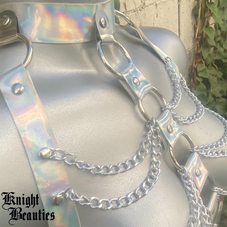 May include: A silver harness with iridescent accents, featuring a choker, shoulder straps, and dangling chains. The harness has metal rings and is secured with rivets. The text "Knight Beauties" is visible at the bottom left.